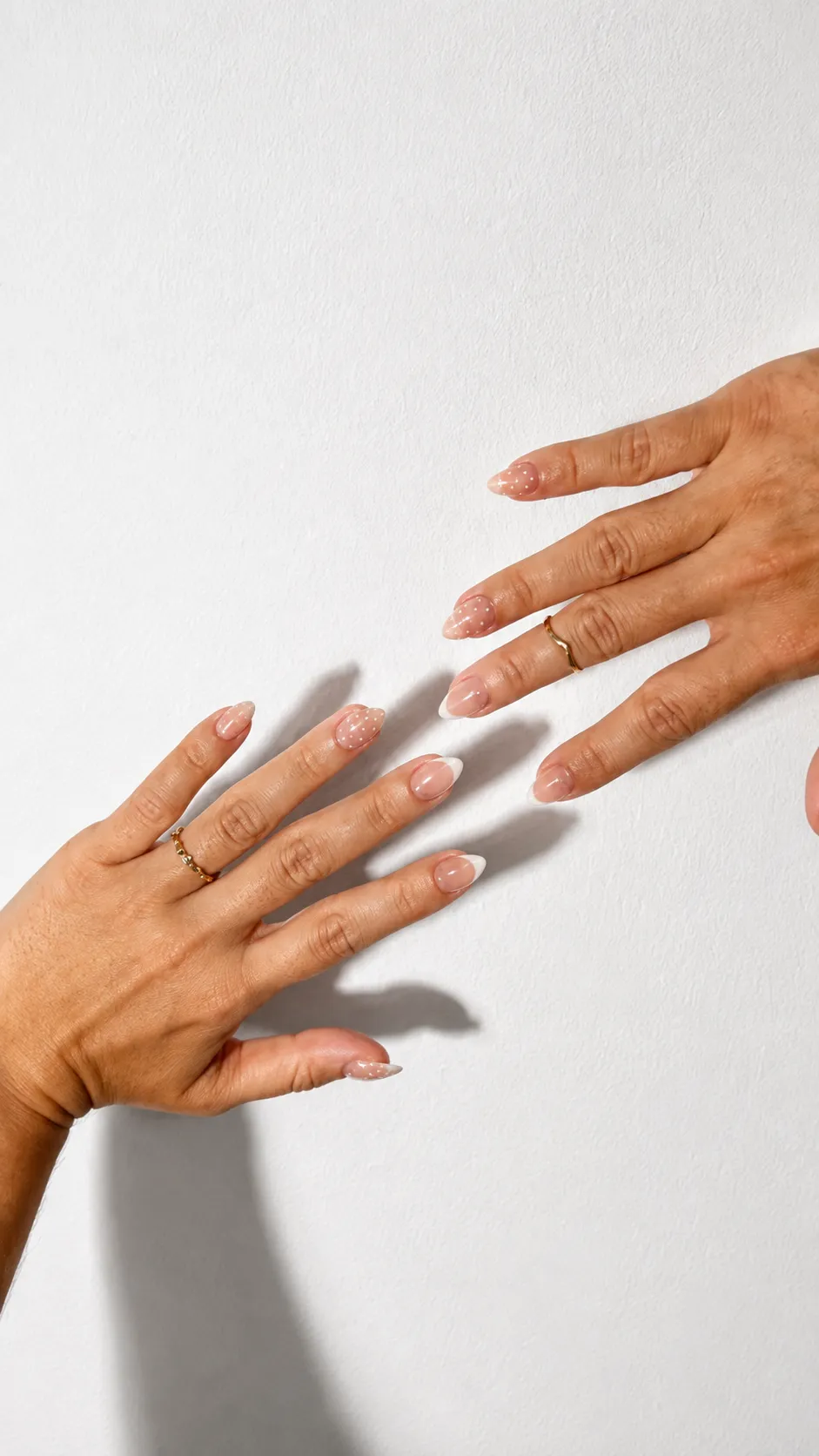 Dos manos sobre fondo blanco con uñas postizas almendra cortas en nude, francesa blanca sutil y lunares blancos