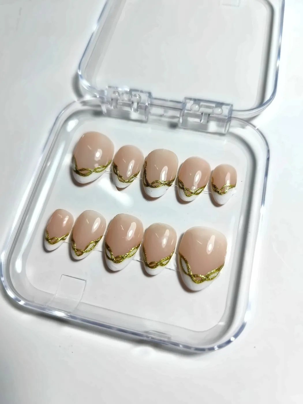 Uñas postizas almendra cortas en nude con francesa blanca y línea dorada ondulada, en su cajita transparente