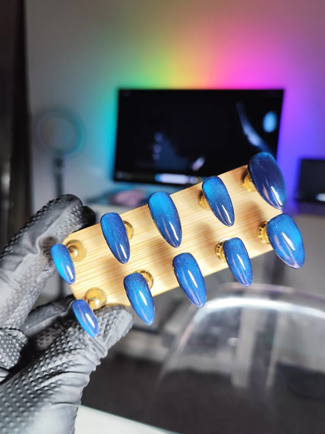 Puntas de uñas postizas almendra en azul eléctrico con efecto ojo de gato, sobre un soporte de madera sujeto con guante negro
