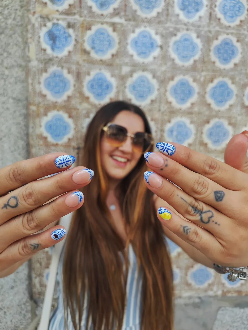 Cristina sonriente, con gafas de sol, mostrando sus uñas inspiradas en azulejo portugués delante de una pared de azulejos azules