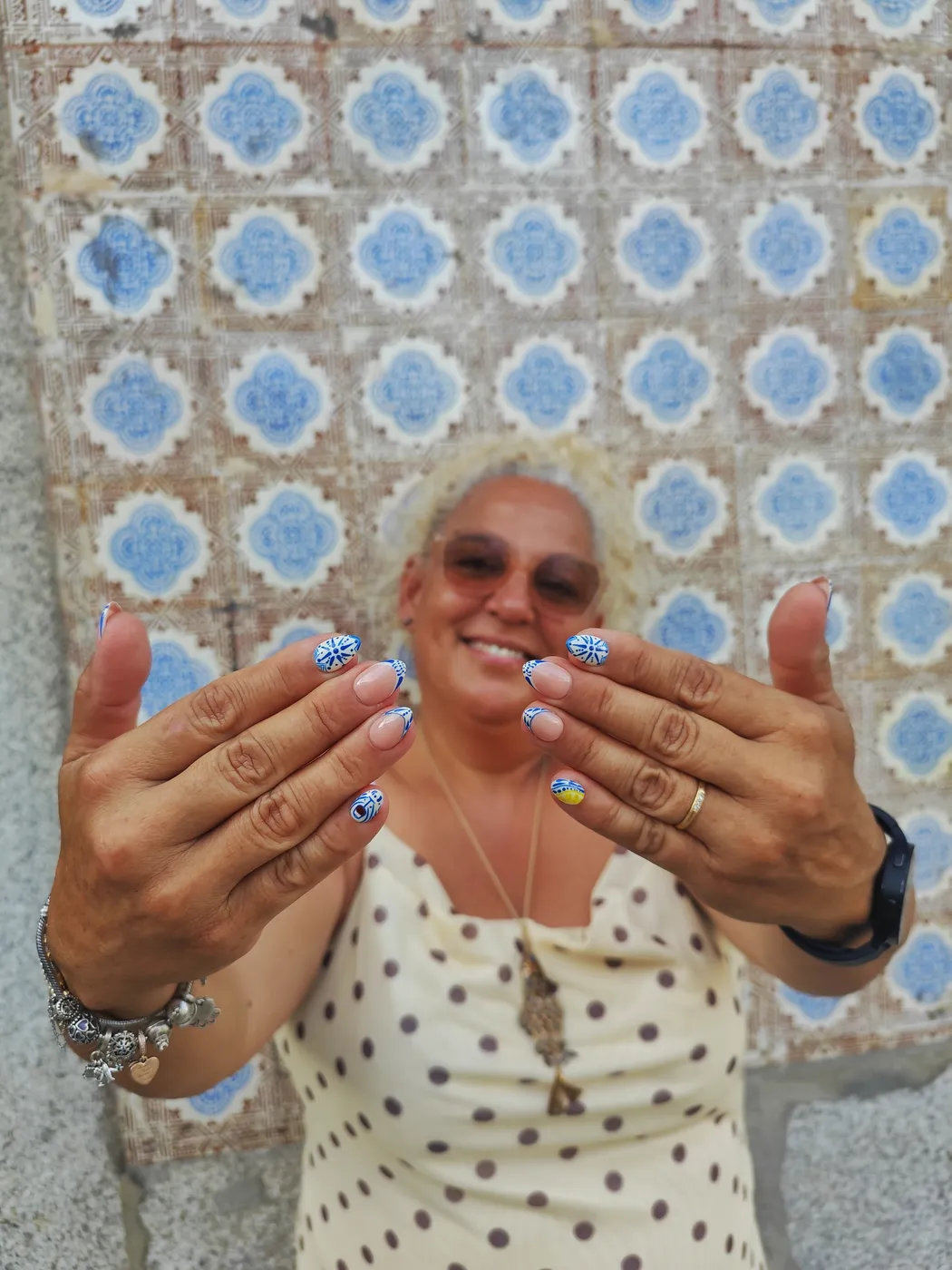 Mujer sonriente mostrando uñas cortas inspiradas en azulejo portugués: dibujos azules sobre blanco, francesa azul y un detalle amarillo, frente a una pared de azulejos