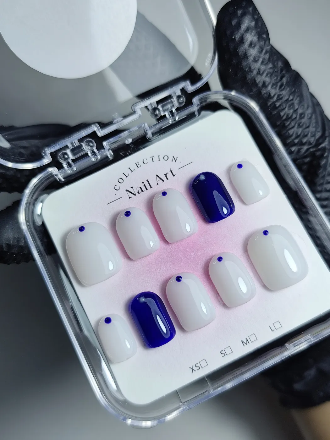 Set de uñas postizas cortas en su estuche, en blanco lechoso con un pequeño punto azul marino en la base y dos uñas en azul marino liso