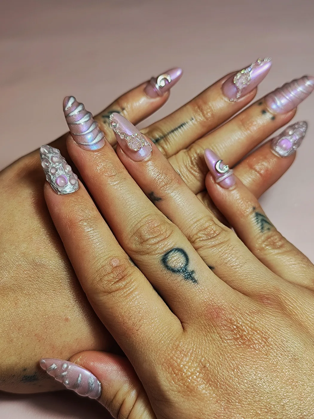 Manos con uñas postizas almendra largas en rosa perlado con efecto cromado, espirales en relieve tipo cuerno de unicornio, charms de luna, perlas y pedrería