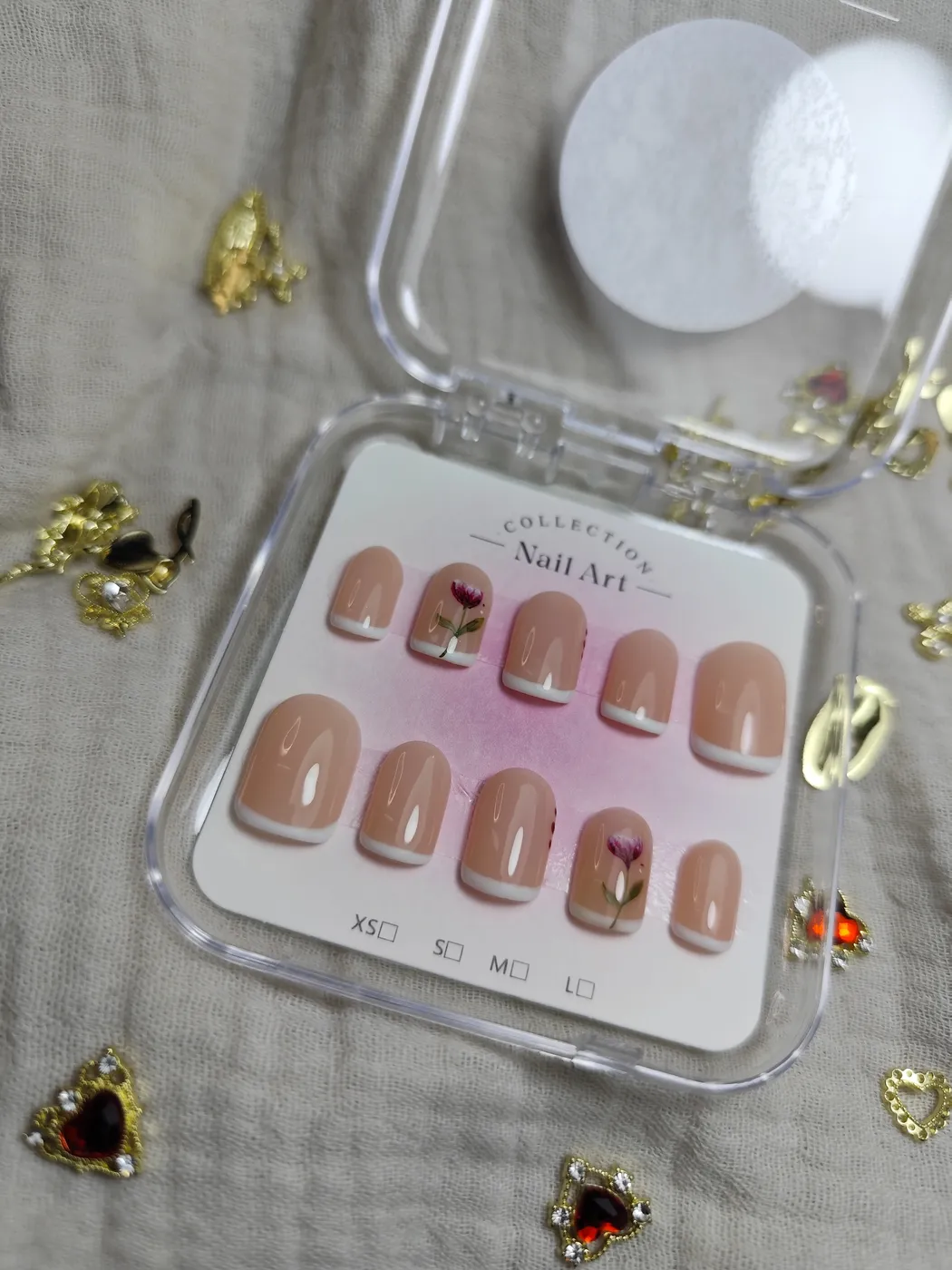 Set de uñas postizas cortas en su estuche, en nude con francesa blanca y dos uñas con un tulipán pintado a mano, rodeado de charms dorados y piedras rojas en forma de corazón