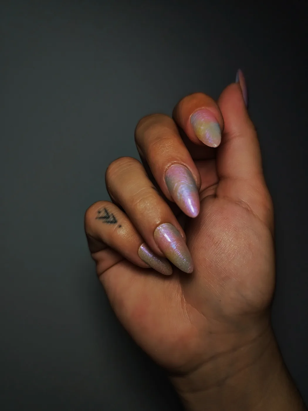 Mano con uñas postizas almendra de efecto iridiscente en tonos pastel rosa, amarillo y azul, sobre fondo oscuro