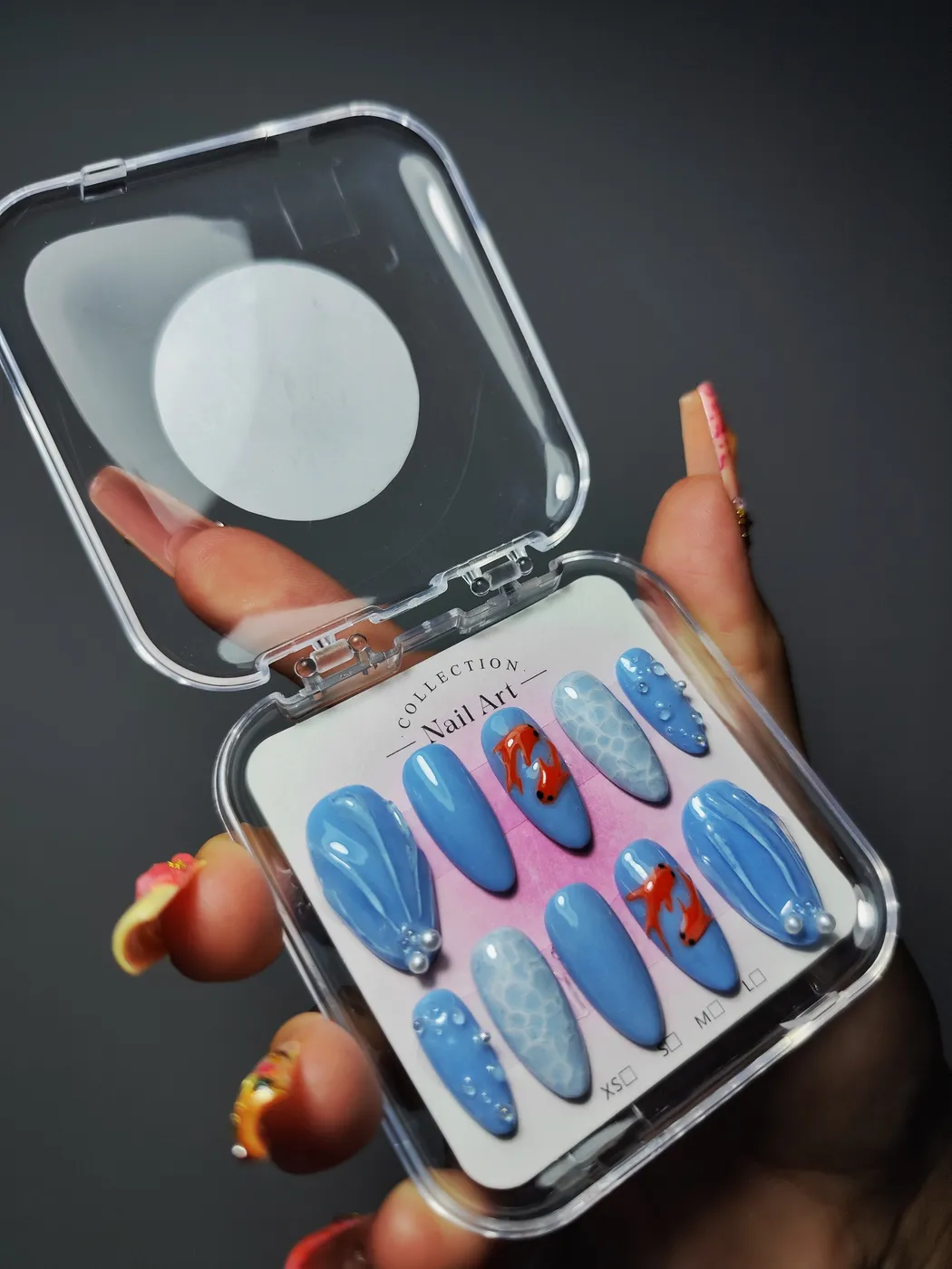 Set de uñas postizas almendra en su estuche en azul cielo con peces koi rojos, efecto de reflejo de agua, ondas en relieve con perlas y gotas de agua