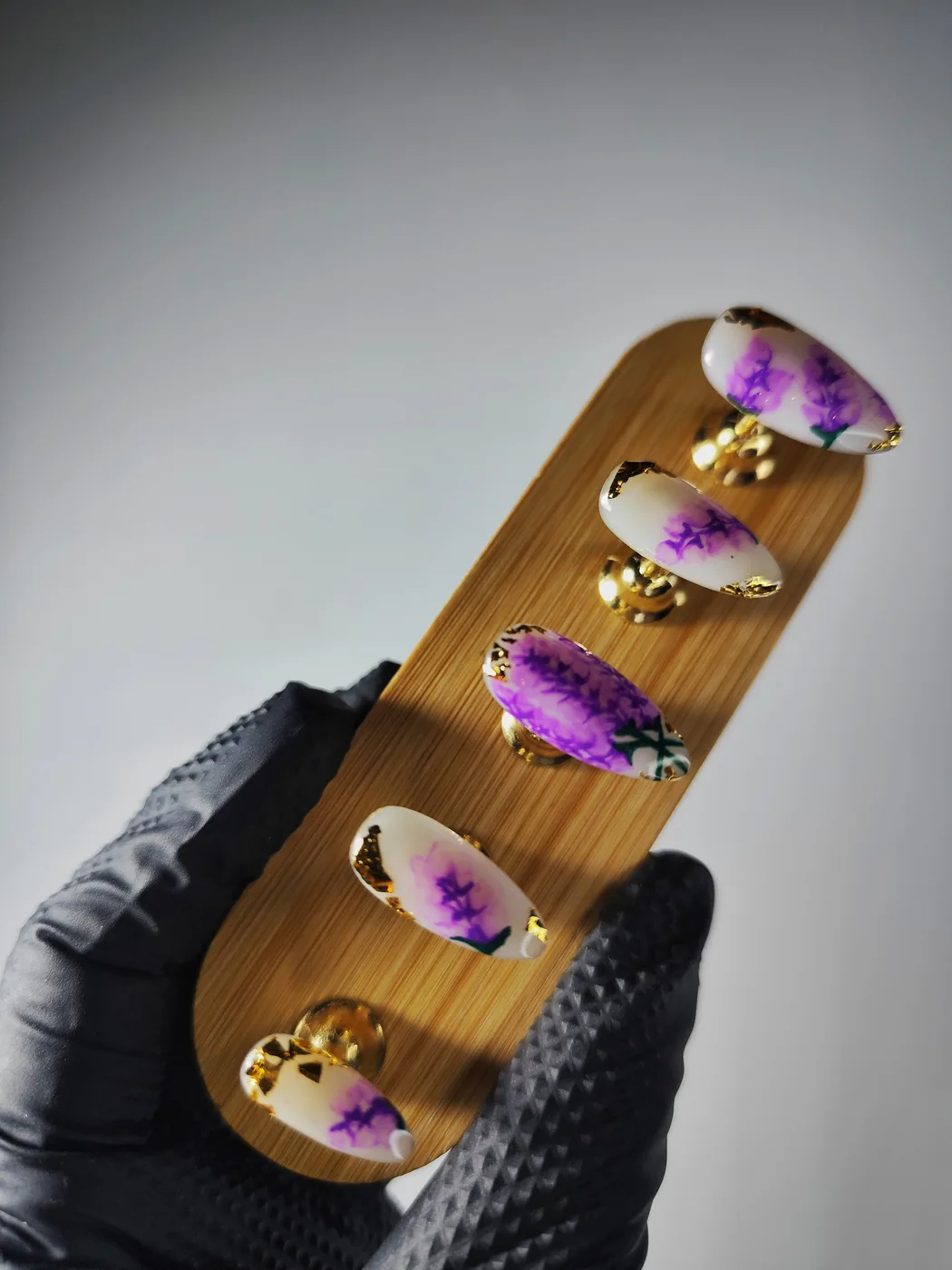 Puntas de uñas postizas sobre un soporte de madera sujeto con guante negro: fondo blanco con lavanda morada pintada a mano y pan de oro