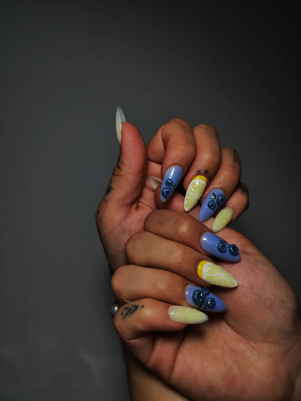 Manos con uñas postizas almendra que alternan amarillo pastel con rodajas de limón pintadas y lila con arándanos en relieve