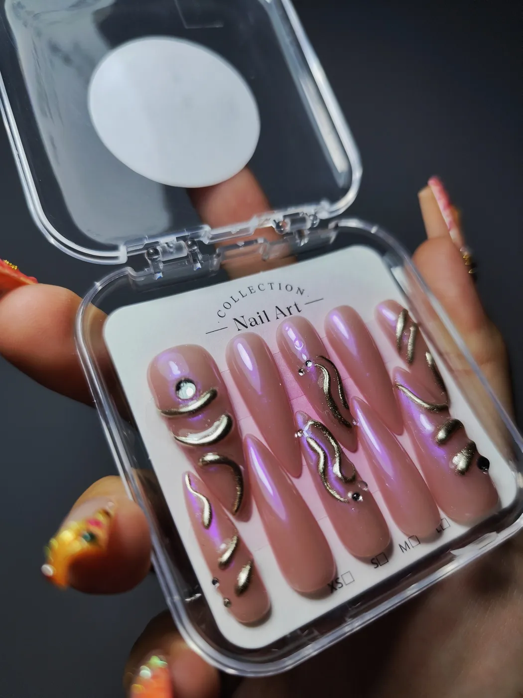 Set de uñas postizas coffin muy largas en su estuche, en rosa nude con reflejo lila, líneas doradas en relieve y pedrería
