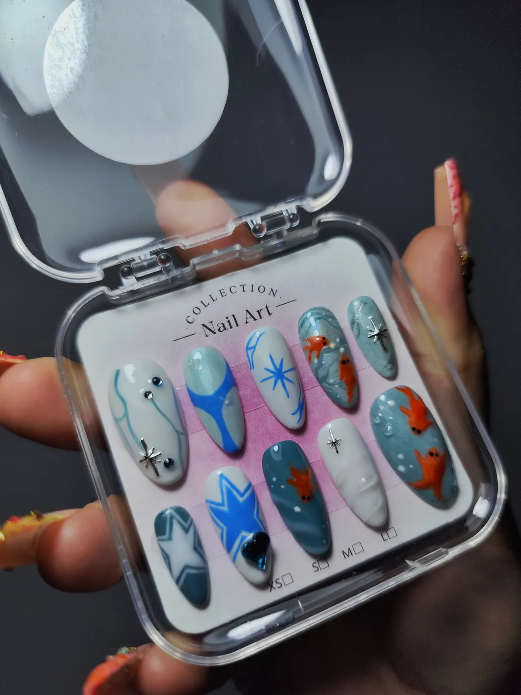 Set de uñas postizas almendra en su estuche de temática marina: peces naranjas pintados a mano sobre agua azul, estrellas y líneas azules sobre blanco, charms de estrella plateados y un strass de corazón azul