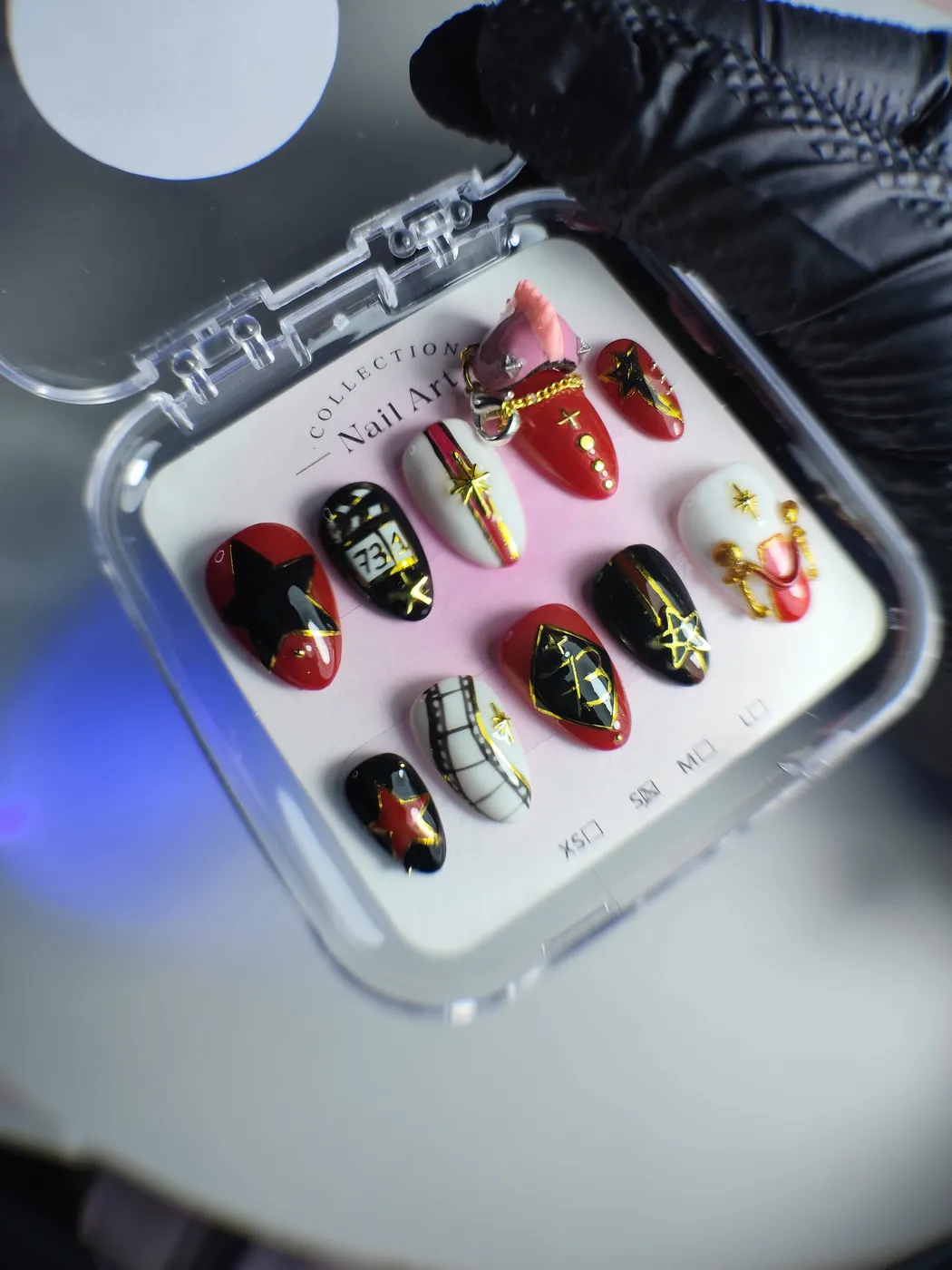Set de uñas postizas en su estuche con diseños combinados en rojo, negro y blanco: estrellas doradas, tira de película, cadenas y un charm rosa en 3D