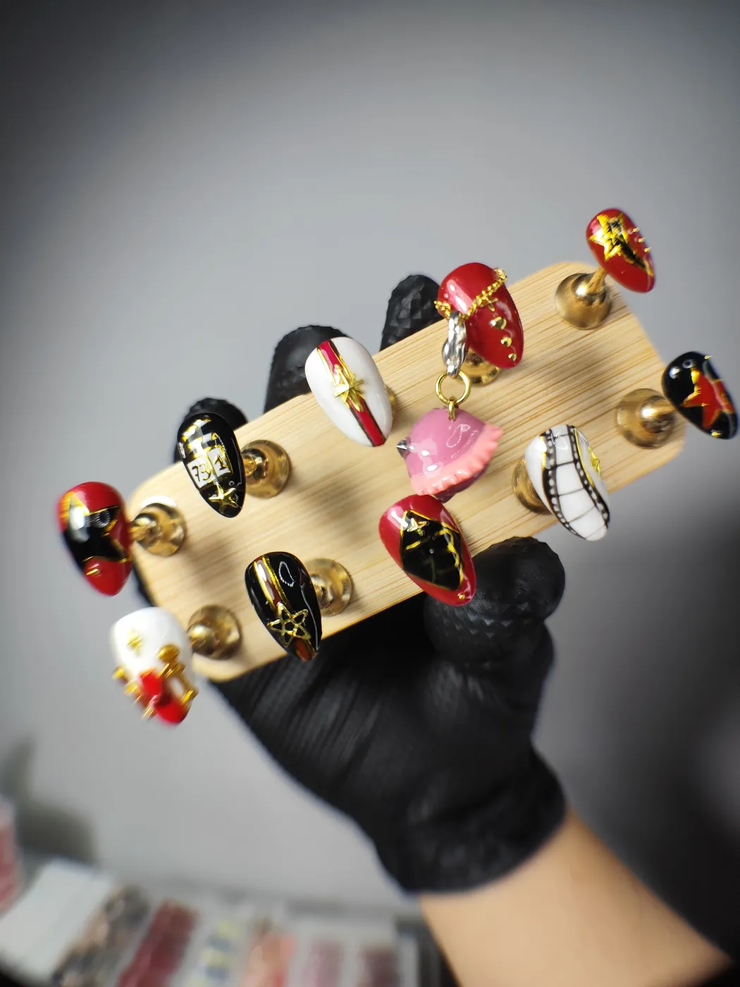 Puntas de uñas postizas sobre un soporte de madera con diseños combinados en rojo, negro y blanco: estrellas doradas, tira de película, cadenas y un charm rosa en 3D