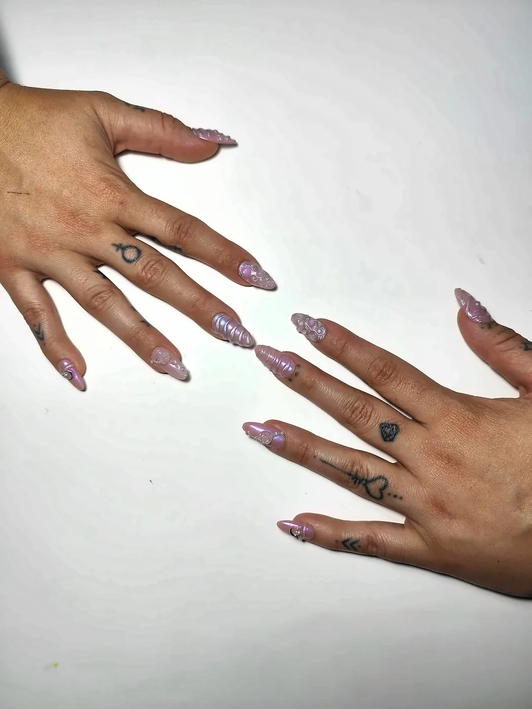 Dos manos abiertas con uñas postizas almendra largas en rosa nacarado con relieve 3D, efecto cromado unicornio y charms