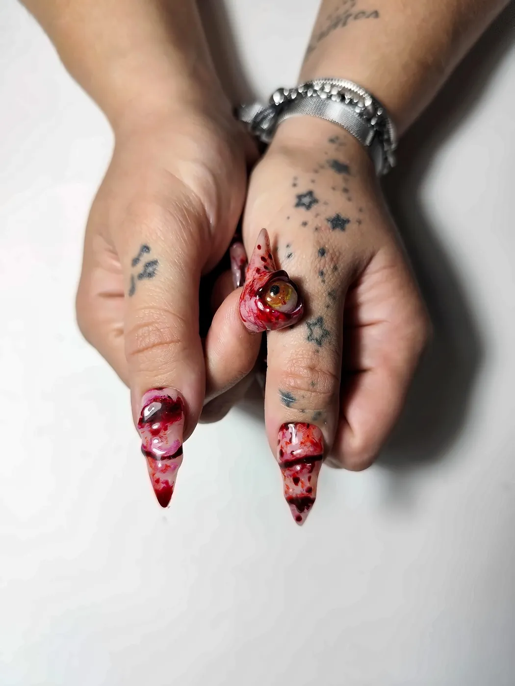 Pulgares con uñas postizas stiletto con efecto sangre y heridas en relieve, junto a un ojo 3D