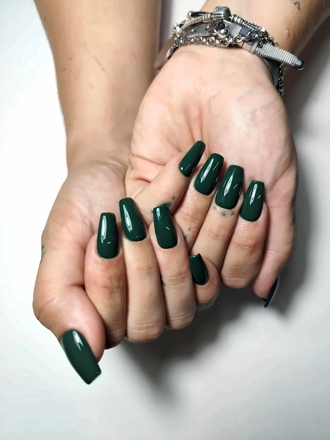 Dos manos con uñas postizas coffin lisas en verde botella, con acabado brillante, una sobre otra